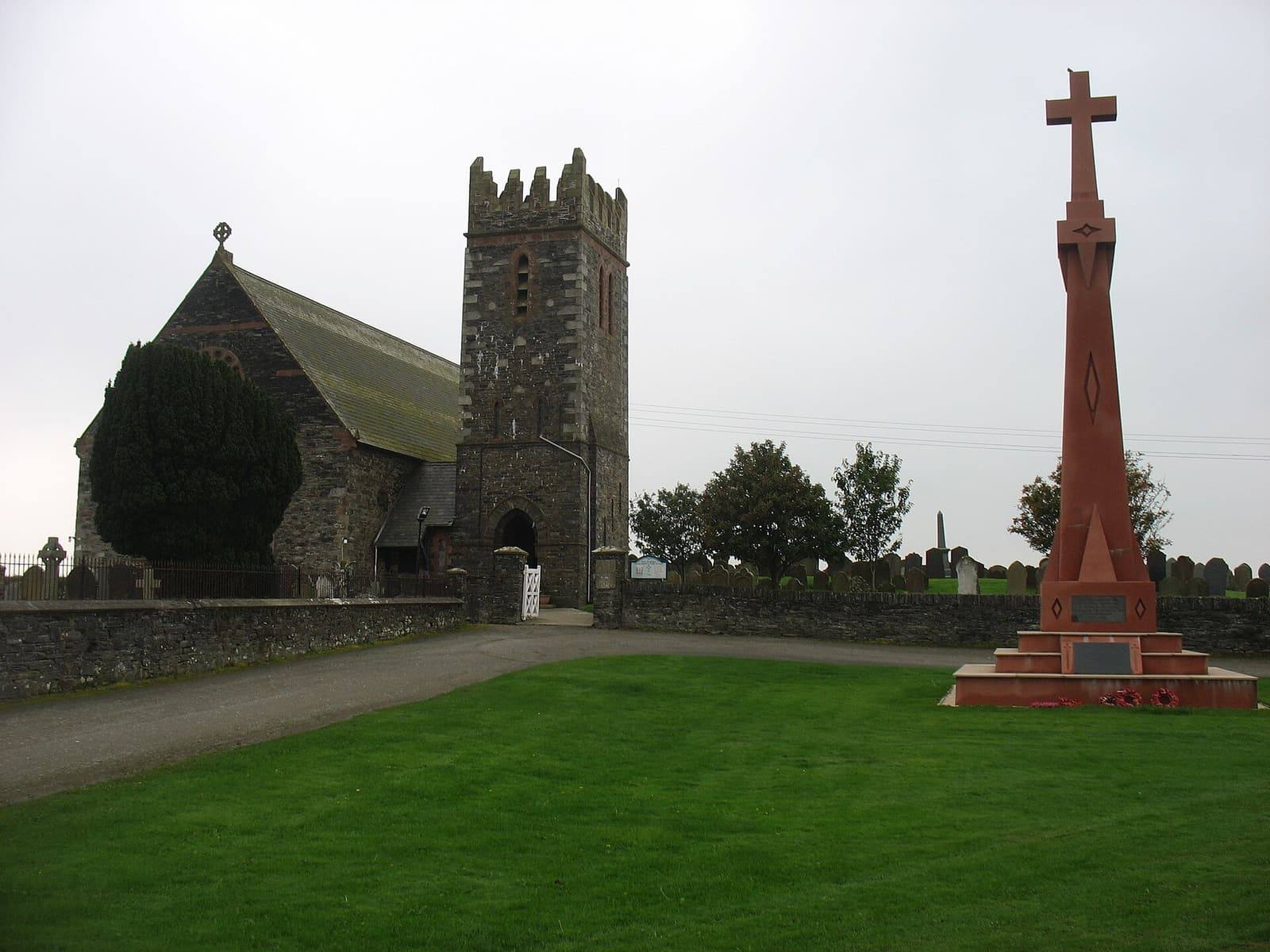 The stone church of St Andrew at Andreas with its square tower, beside a tall red sandstone war memorial cross.
