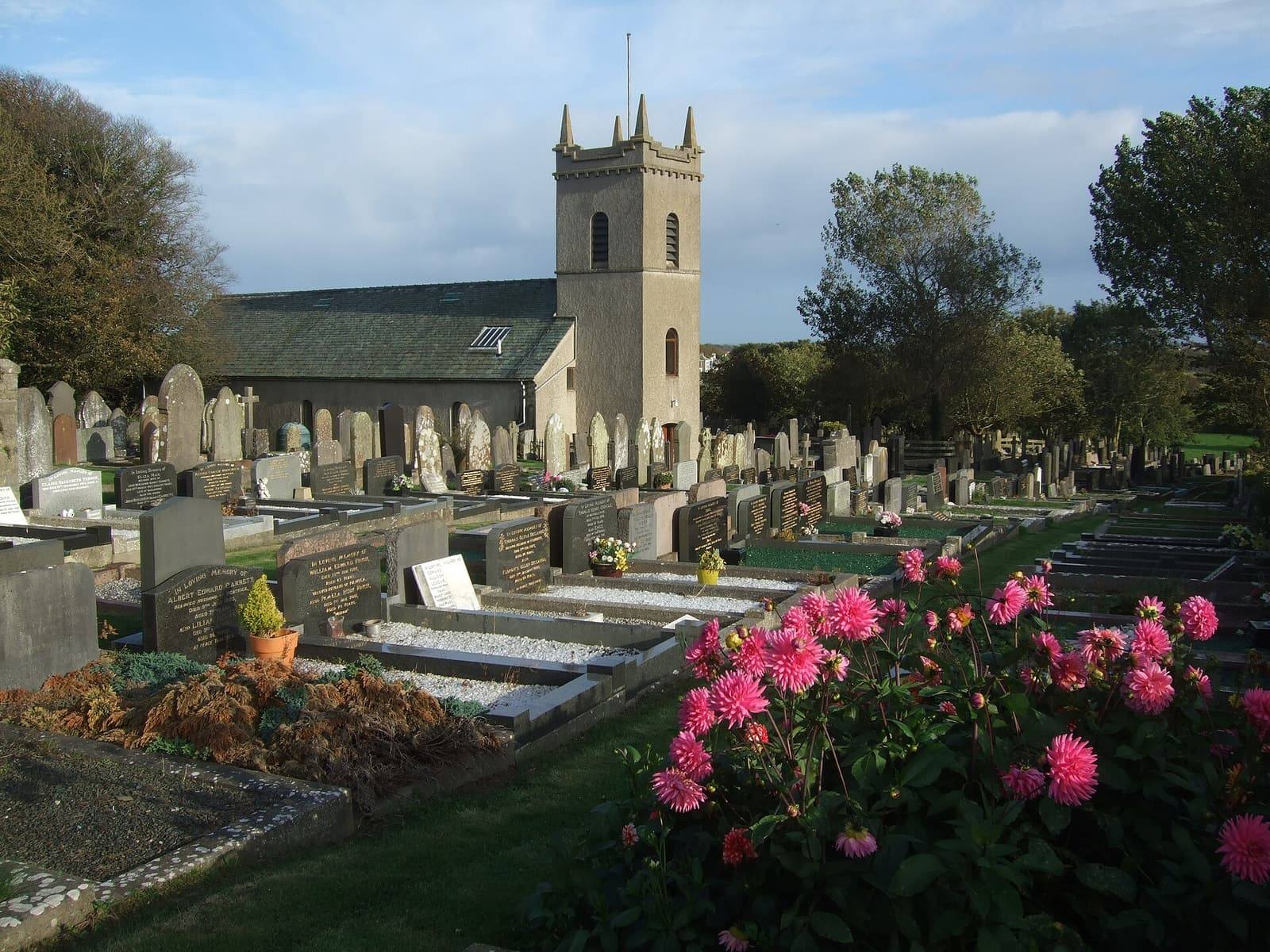 The stone parish church of Arbory at Ballabeg with its square tower, pink flowers and gravestones in the churchyard in low autumn sun.