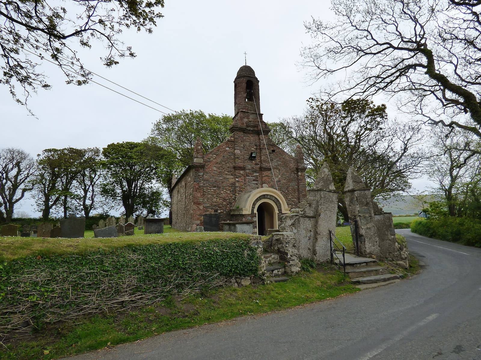 The small stone Old Church at Ballaugh with its bell-cote, behind a hedge on a quiet country road.