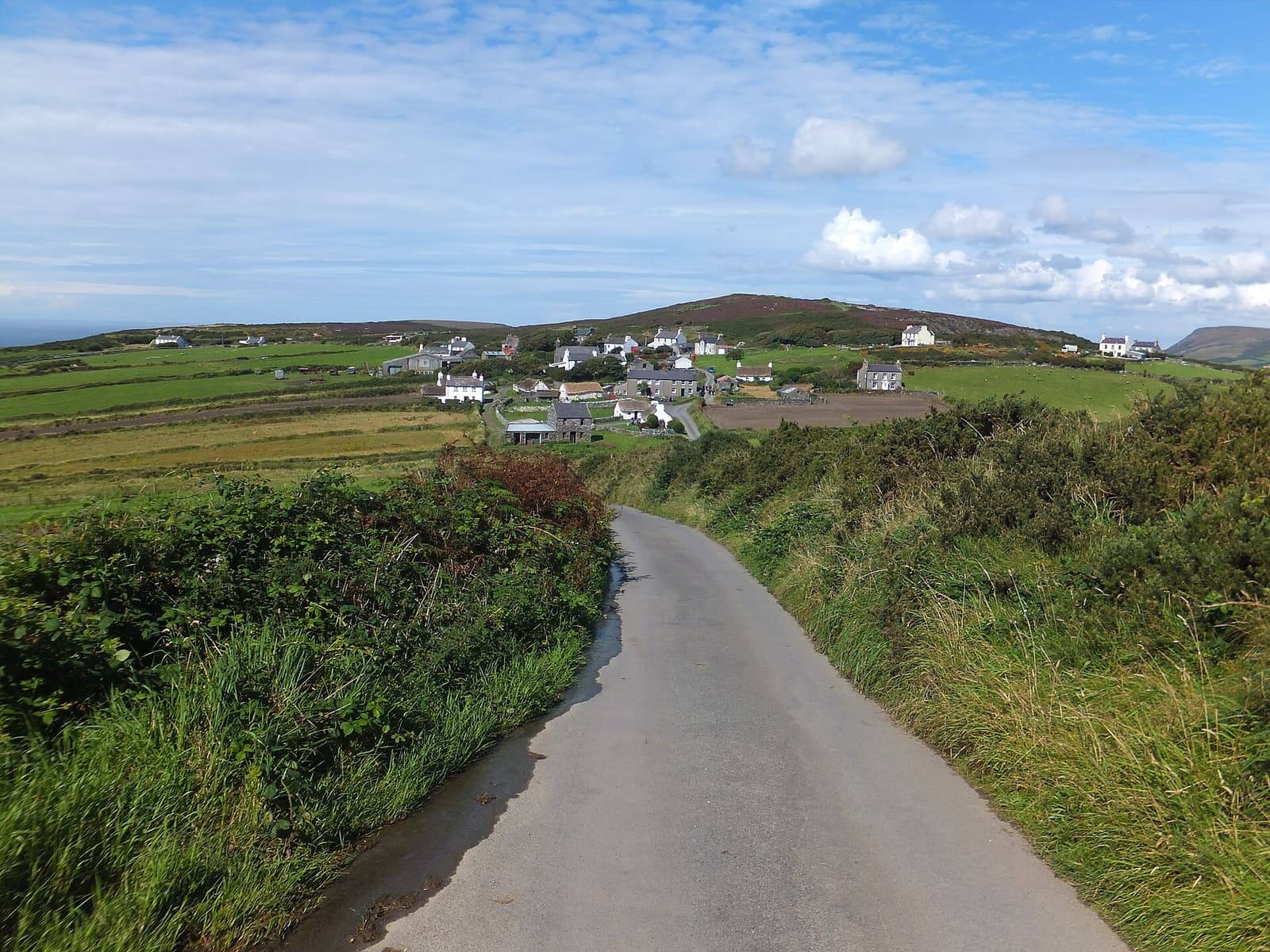 A narrow lane between green banks leading up to the white cottages of Cregneash on the hill under a blue sky.