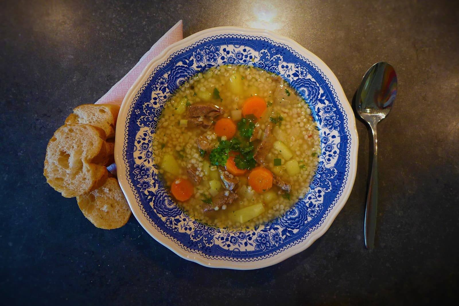 A blue-and-white bowl of barley soup with beef, carrot, potato and parsley, with bread and a spoon beside it.