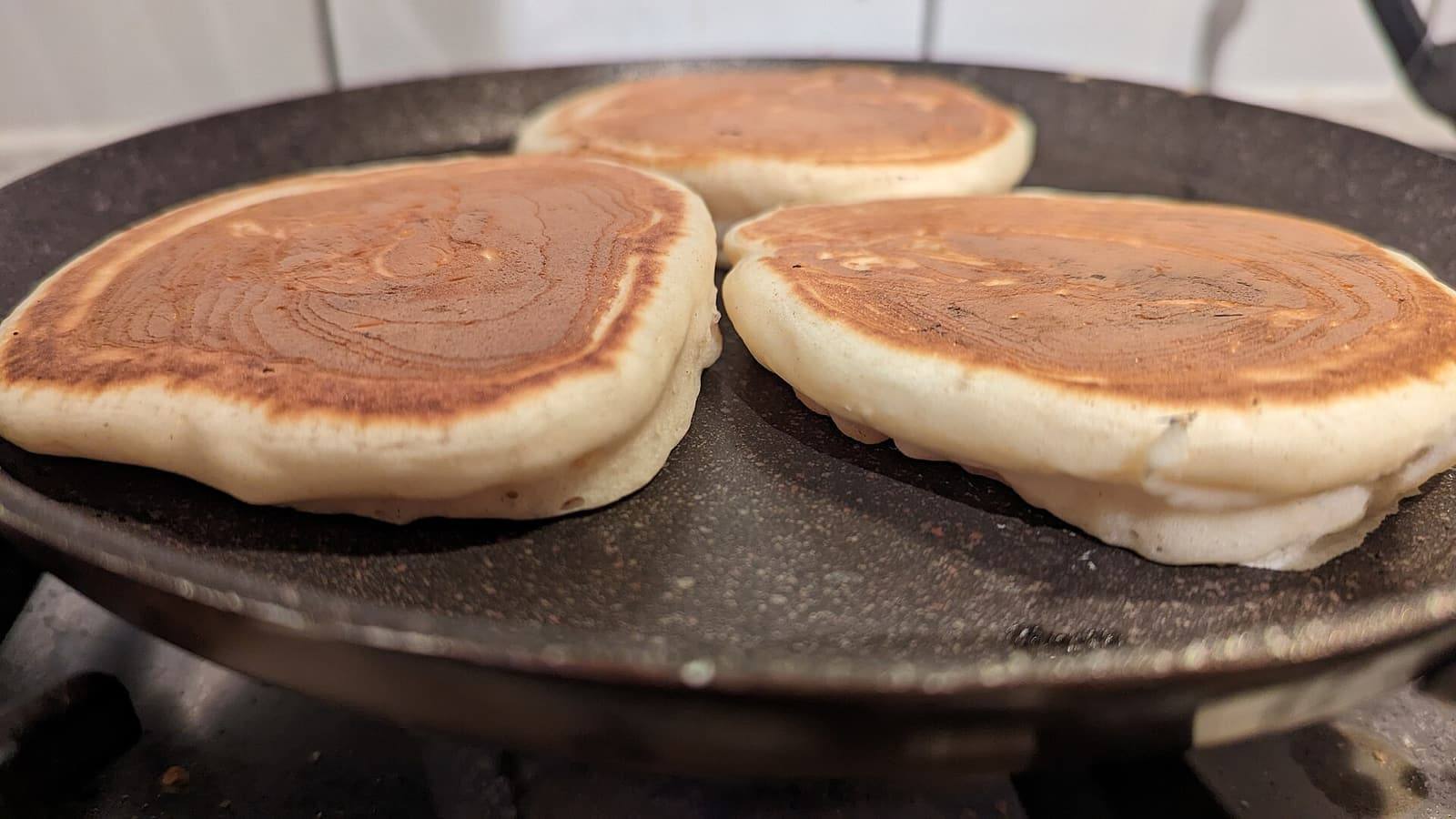 Three thick, golden buttermilk pancakes cooking on a round black griddle.