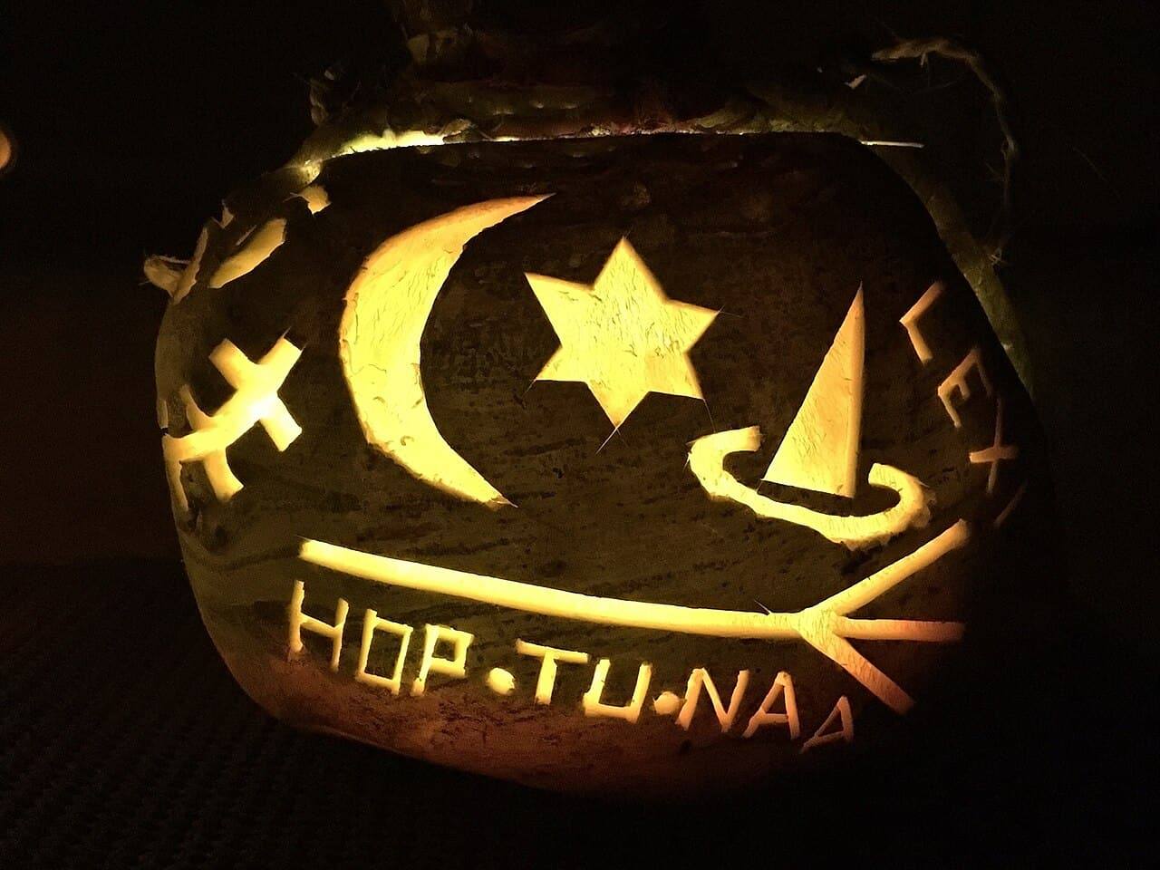 A turnip lantern glowing in the dark, carved with a moon, a star, a witch's hat and the words Hop-tu-naa.