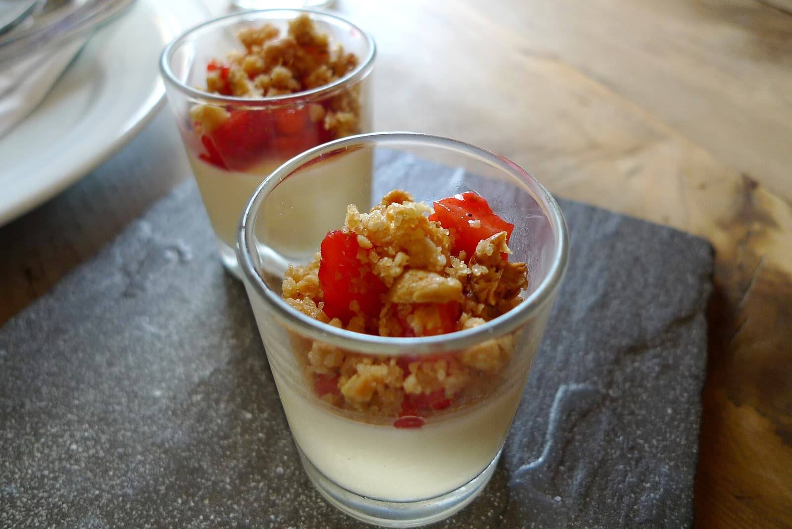 Two small glasses of pale set junket topped with chopped strawberries and crumble on a slate.
