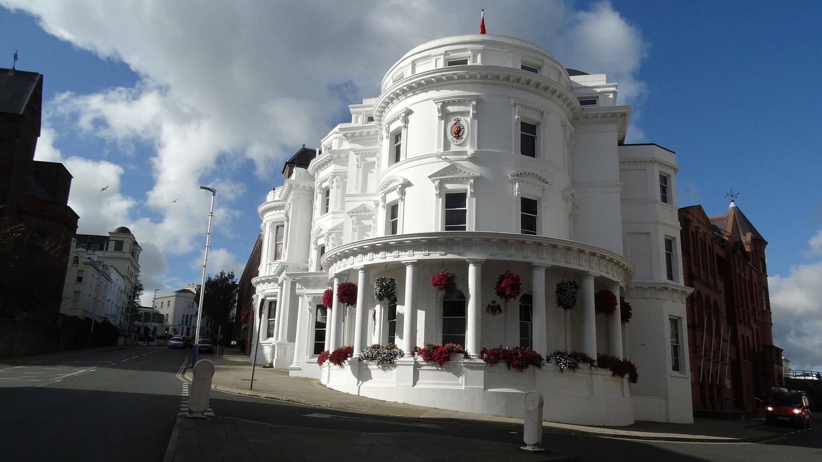 The white, curved, colonnaded government building on the corner of Prospect Hill in Douglas under a blue sky.