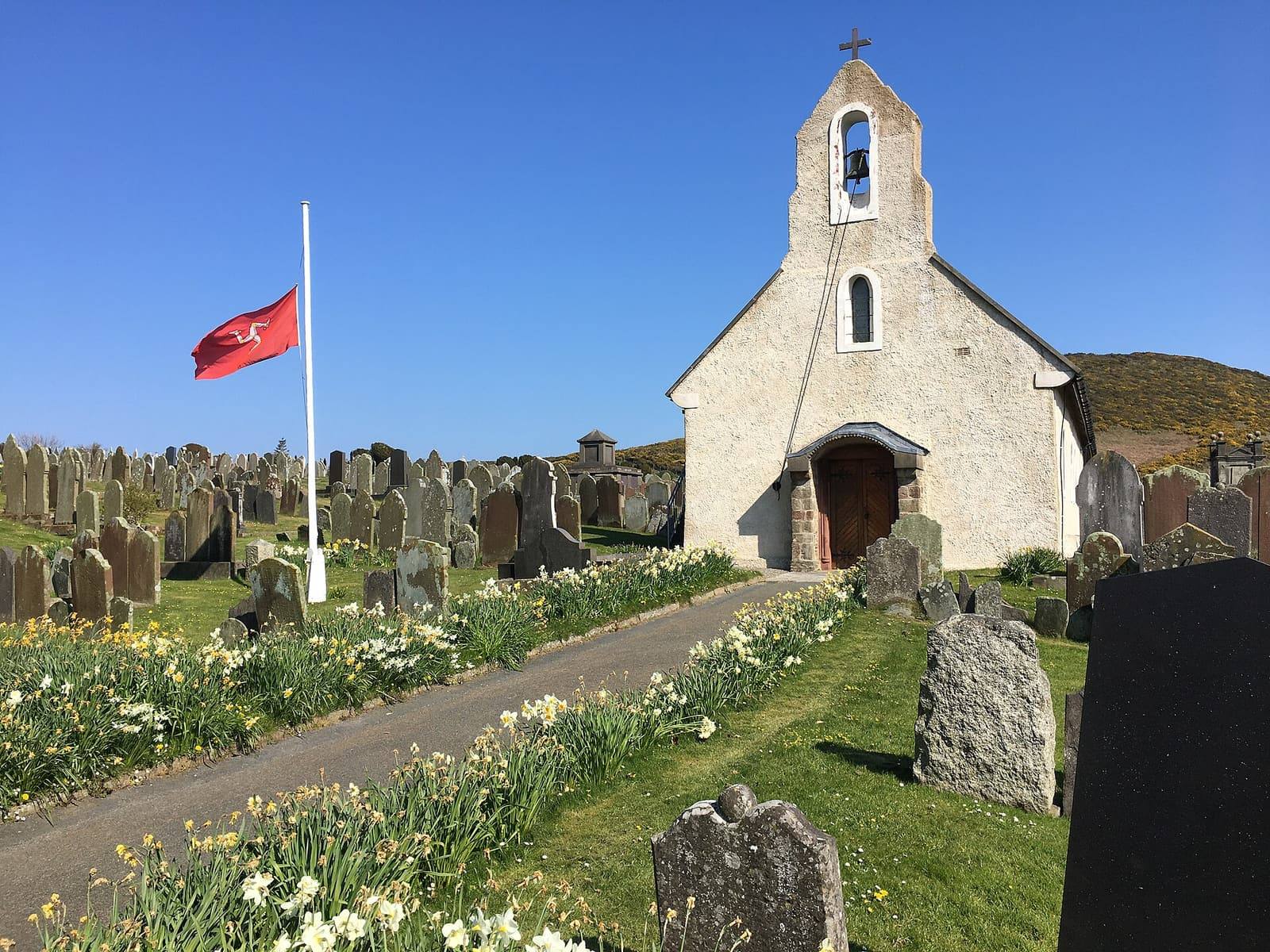 The whitewashed Kirk Maughold church with a Manx flag flying beside it, daffodils along the path between old gravestones.