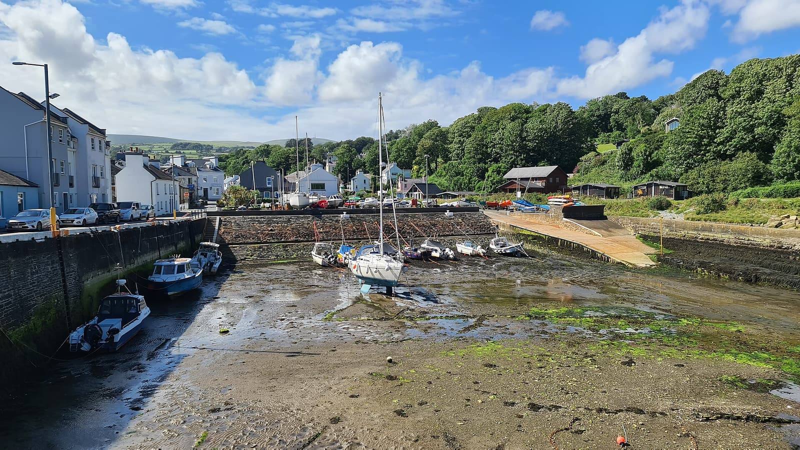 Small boats on the mud of Laxey's little harbour, with white cottages and wooded hills behind.
