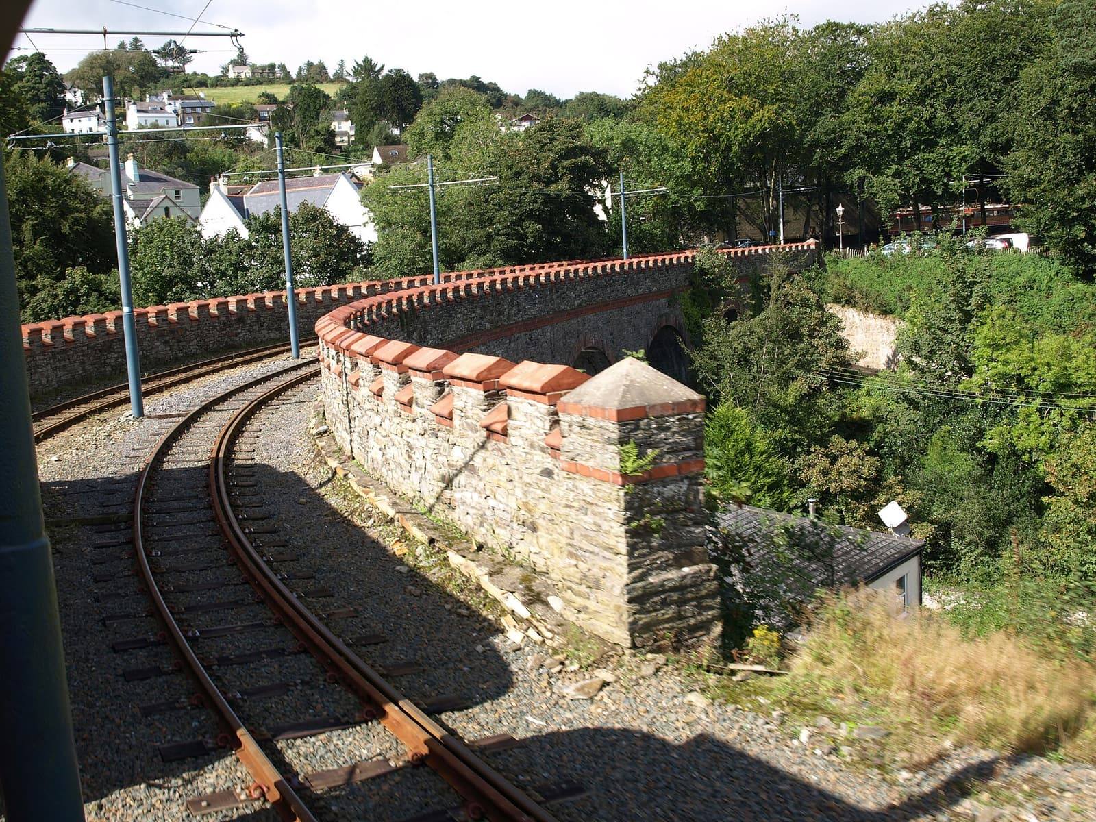 The tram line curving along a stone embankment into Laxey, with wooded hillside and cottages below.