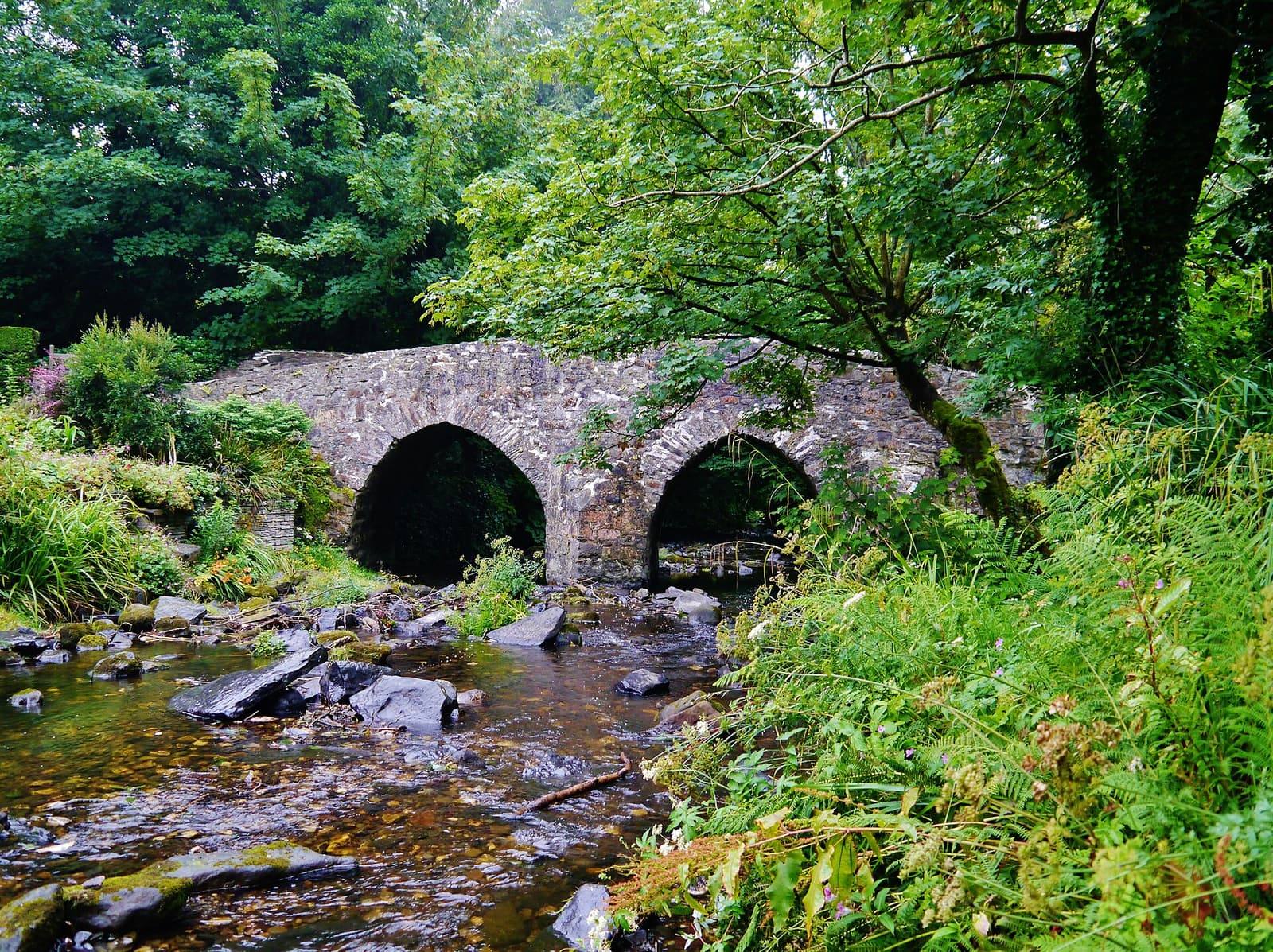 The old stone arches of Monks' Bridge over the Silverburn river at Ballasalla, surrounded by green ferns and trees.