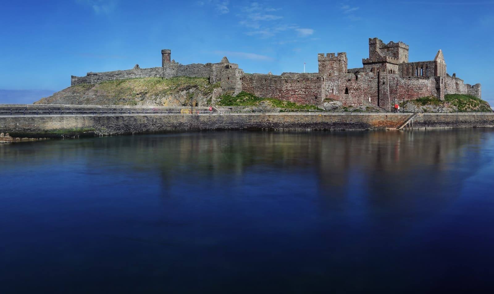 Peel Castle's long red sandstone walls reflected in calm blue water.