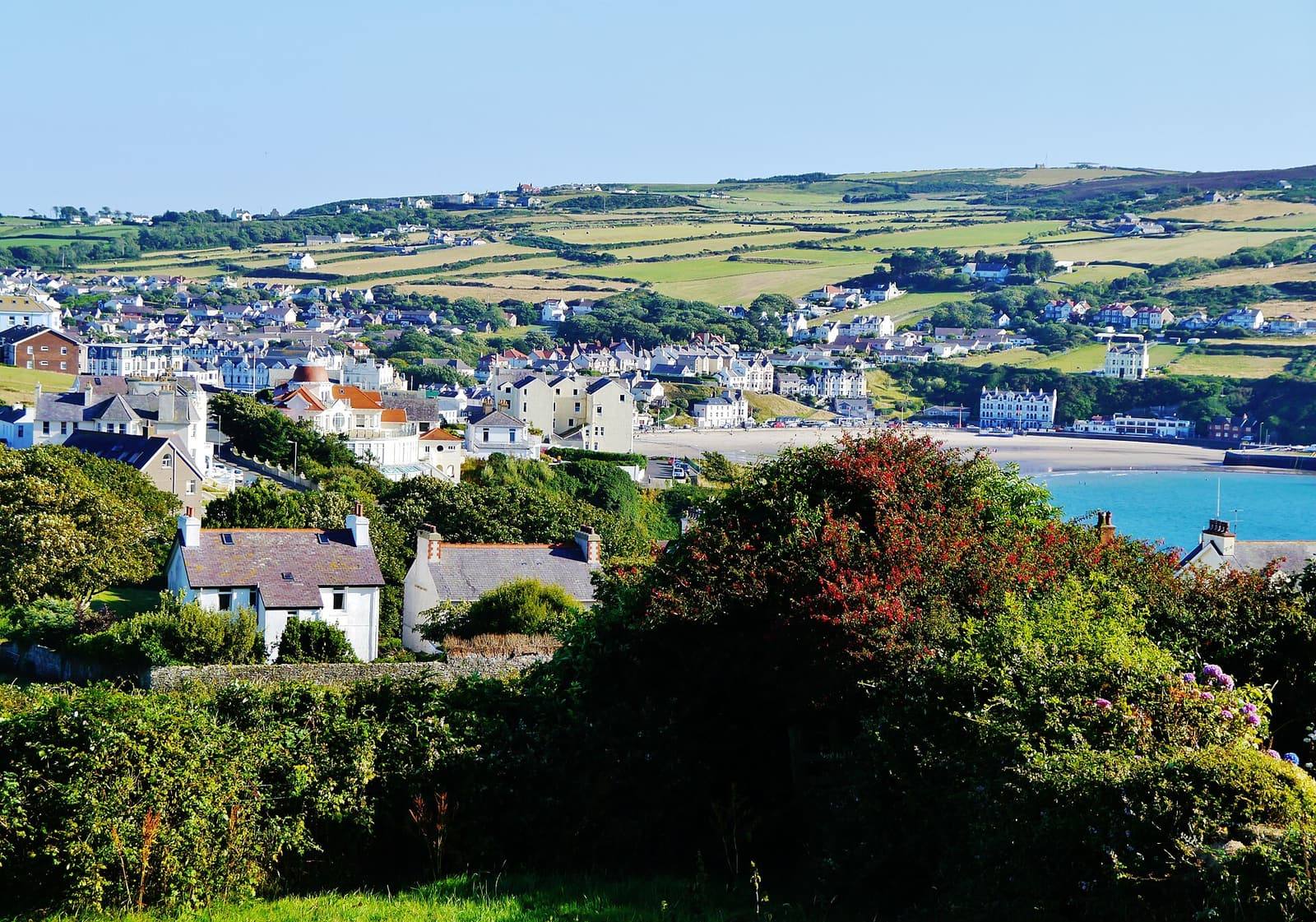 Port Erin's houses and sandy bay in sunshine, seen across gardens and trees, with green fields rising behind.