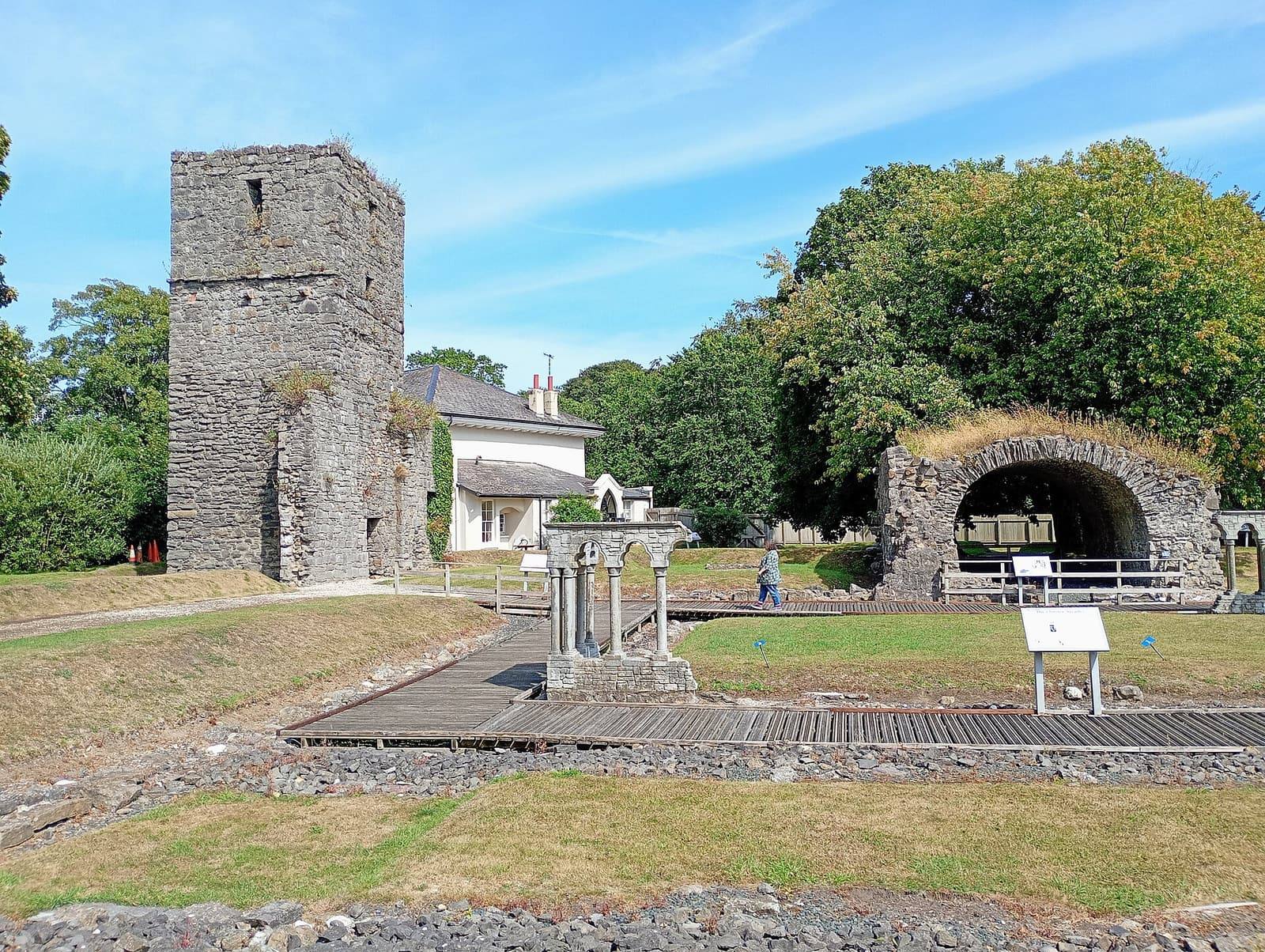 The stone tower and ruined walls of Rushen Abbey at Ballasalla on a sunny day, with trees and lawns around them.