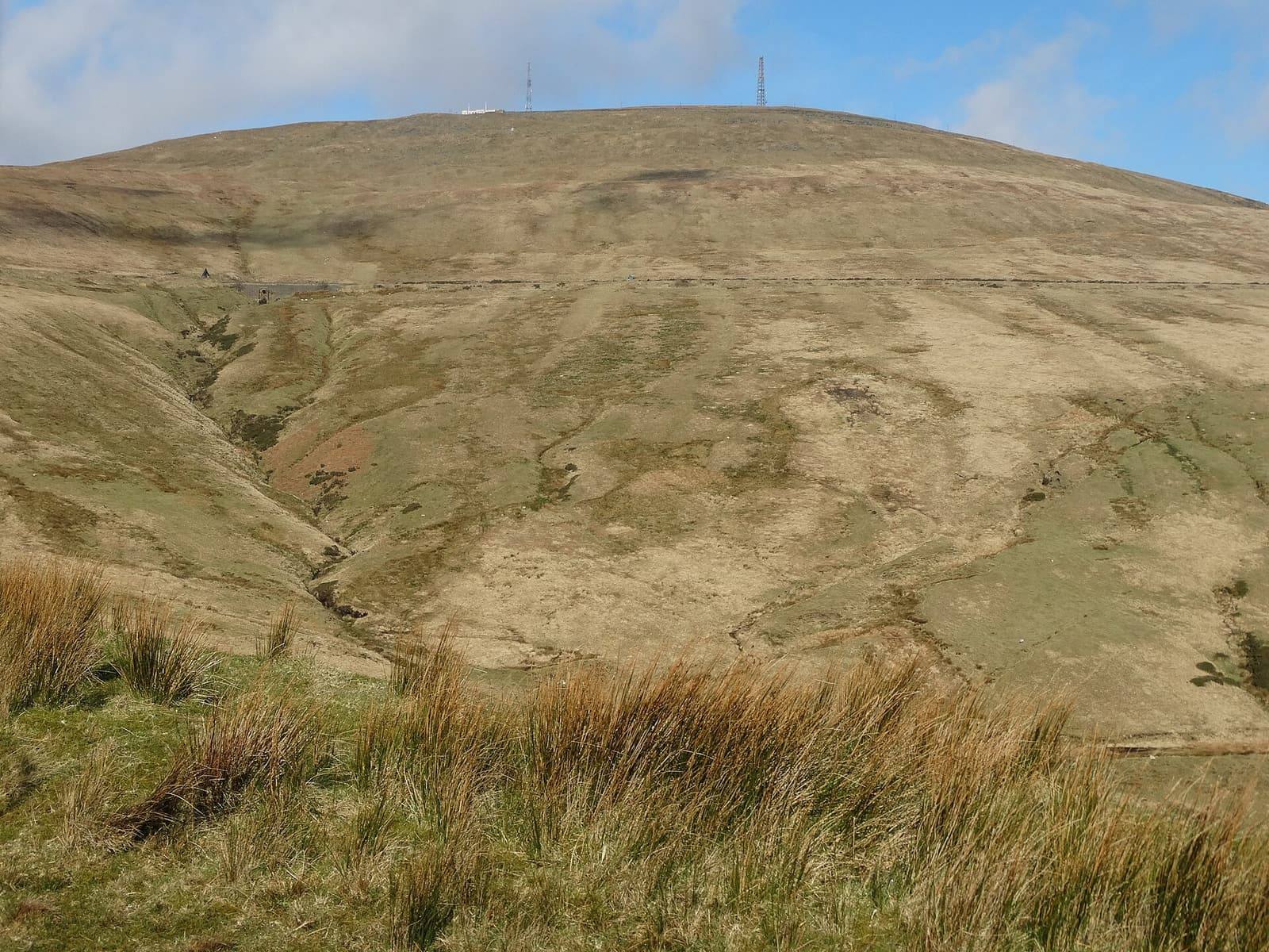 Open brown and green moorland rolling up towards the summit of Snaefell under a wide blue sky.