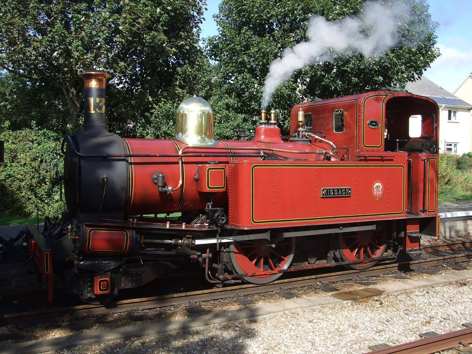 A red Isle of Man Railway steam engine with a brass dome letting off steam at Colby station among trees.