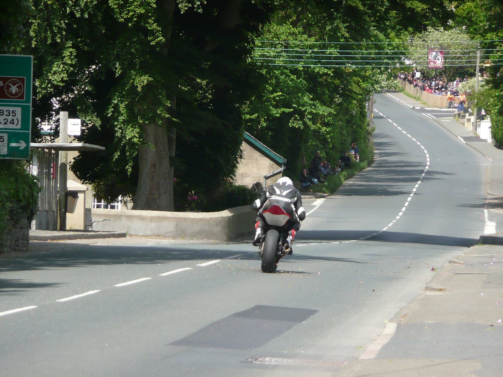 A motorcycle racer approaching Crosby crossroads between tall trees during a TT race, with spectators far down the road.